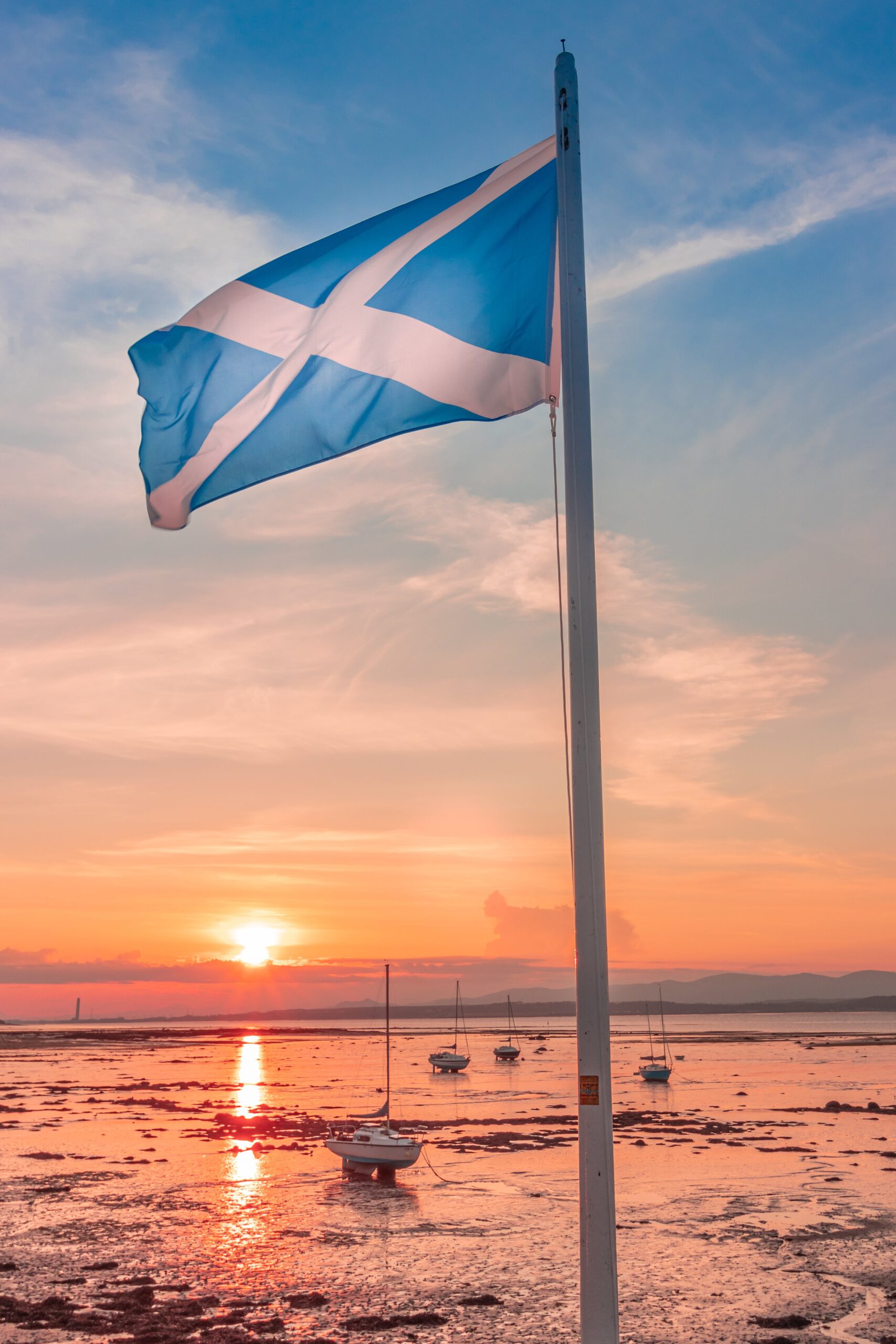 The Scottish flag on a flagpole with a beach at sunset in the background.