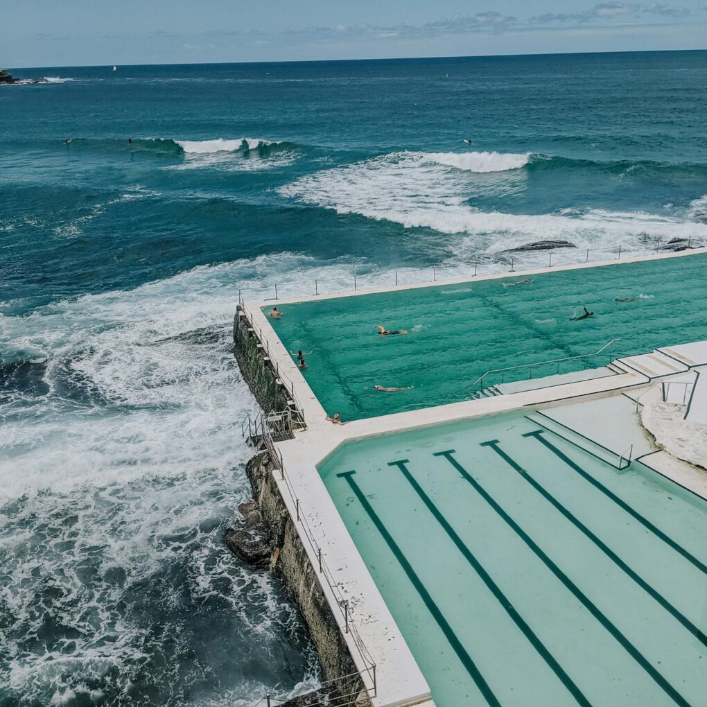 Ocean pool beside crashing waves, with swimmers and surfers in the sea beyond.