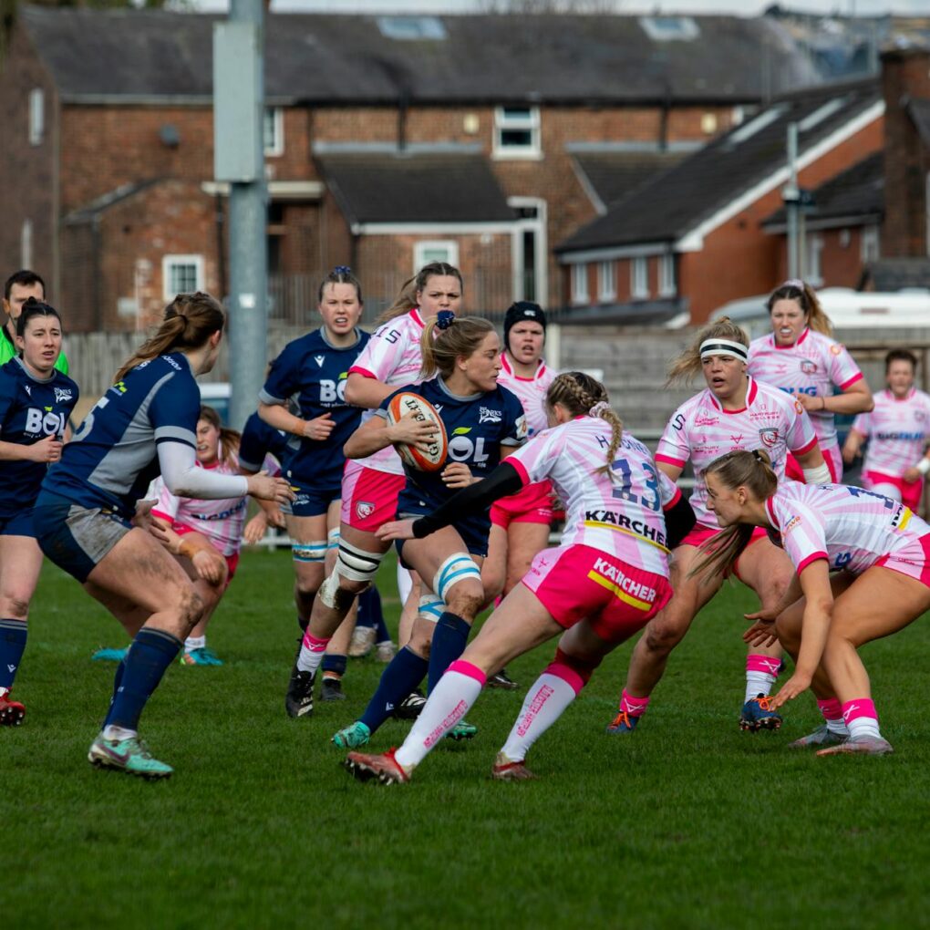 Two teams of female rugby players on a pitch playing a match.