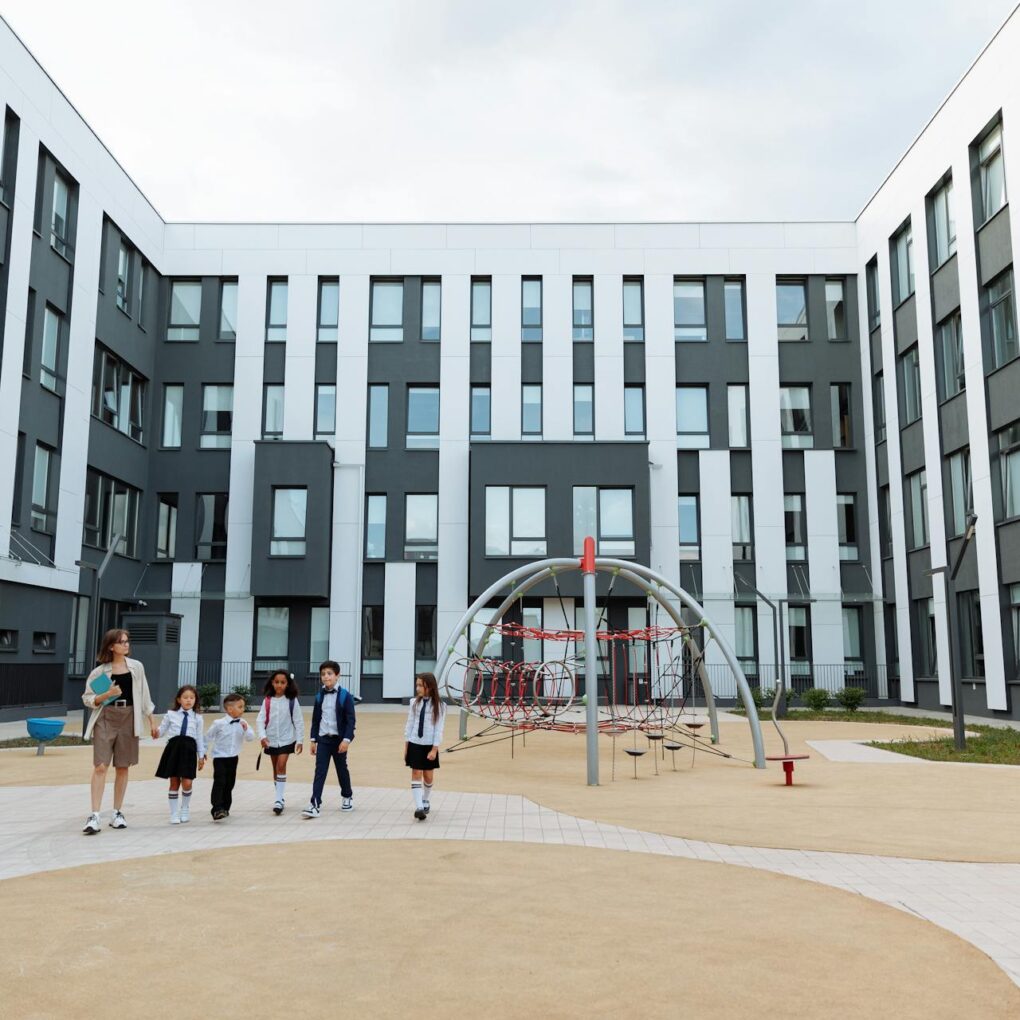 Children and a teacher walking through a modern school courtyard with contemporary playground equipment.