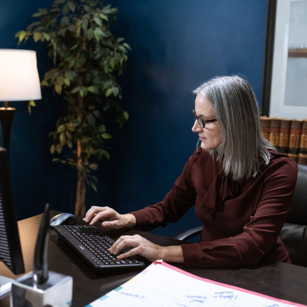 Woman with grey hair in a corporate office typing on a keyboard, focused and professional.