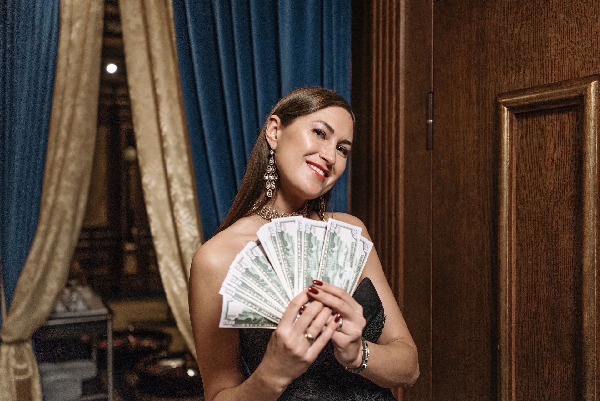 Woman in a black evening dress holding a fan of US dollar bills and smiling at the camera.