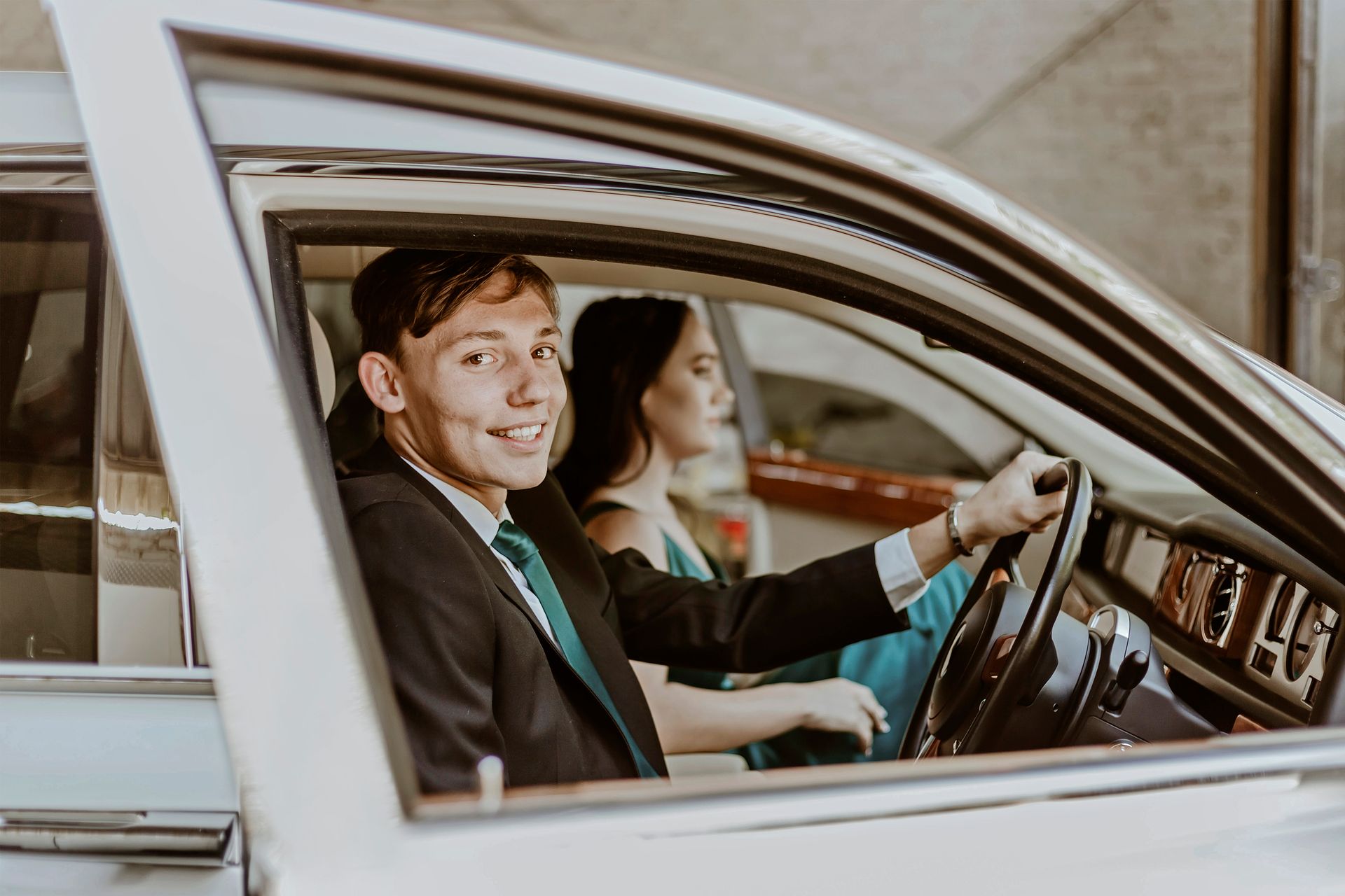 Young man in a suit and green tie smiling from the driver's seat of a car, with a woman sitting beside him.